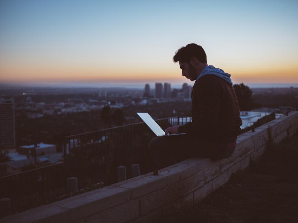 A man working on a laptop while sitting on a rooftop ledge at dusk, with a city behind him
