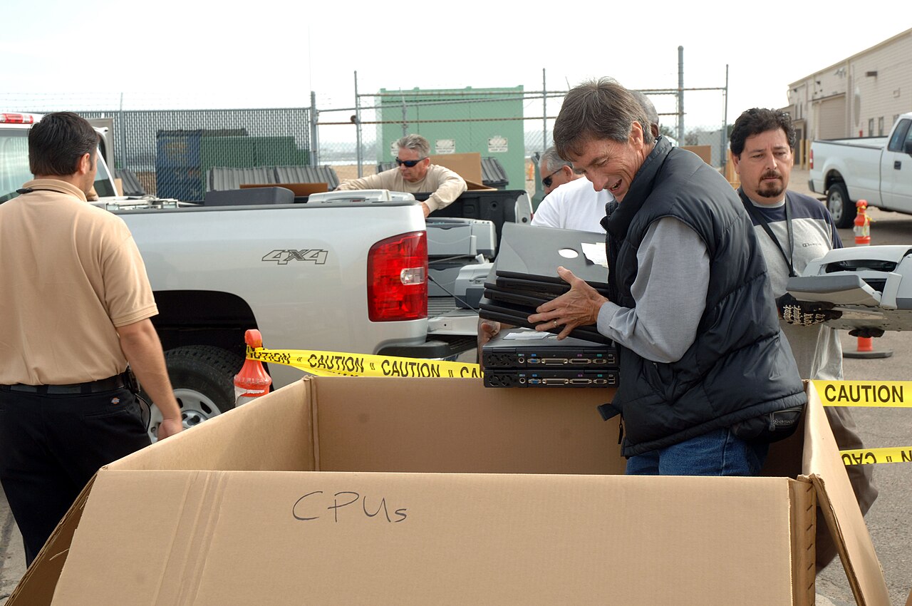 A man loads old computers into a cardboard box labeled CPUs near a pickup truck behind caution tape.